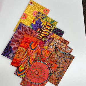 May include: A collection of rectangular fabric swatches arranged in a layered stack. The textiles feature vibrant, multi-coloured patterns including floral, geometric, and abstract designs in shades of orange, yellow, purple, pink, and green.