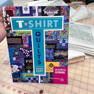 May include: A book titled "T-Shirt Quilts Made Easy" by Martha Deleonardis. The cover features a patchwork design with various fabric squares and the text "T-SHIRT QUILTS" in a teal banner. The book is on a cutting mat.
