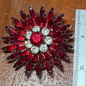 Könnte beinhalten: Eine kreisförmige Brosche mit einem zentralen roten Strassstein, umgeben von einem Ring aus klaren Strasssteinen und mehreren Schichten spitzer, marquise-geschliffener roter Strasssteine. Ein Metalllineal zeigt eine Breite von ca. 5 cm.
