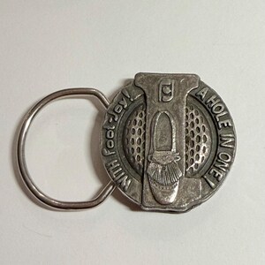 May include: A silver-toned metal keychain featuring a circular medallion with a textured golf ball pattern. The medallion displays a raised golf shoe and the text "WITH FOOT-JOY! A HOLE IN ONE!" along the edge, attached to a round metal ring.