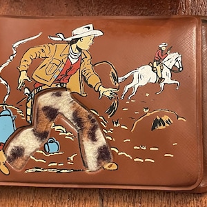Könnte beinhalten: Braune Geldbörse mit einer Illustration zum Thema Cowboy. Das Kunstwerk zeigt einen Cowboy in einer beigefarbenen Jacke und einer Hose mit Muster, der mit einem weißen Pferd im Hintergrund rennt. Eine blaue Kaffeekanne ist ebenfalls zu sehen.