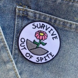 Puede incluir: Parche blanco bordado con una flor rosa que crece de un montículo de tierra gris. El parche dice "Survive Out of Spite" en un borde circular negro.