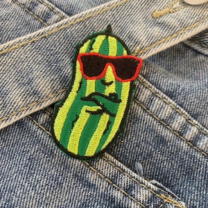 May include: An embroidered patch of a cucumber wearing sunglasses. The cucumber has a green and yellow striped pattern and a red mouth. The sunglasses are black with red frames.