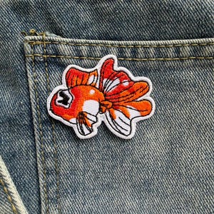 Puede incluir: Parche bordado con un diseño de pez dorado en tonos naranja, blanco y negro, aplicado en el bolsillo de unos vaqueros azules. El parche presenta un diseño detallado de pez dorado con aletas fluidas y una estética lúdica.
