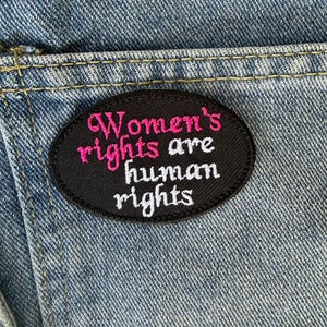 Op de afbeelding: Een ovaal zwart embleem geborduurd met de woorden "Women's rights are human rights" in roze en witte draad. De patch is genaaid op een blauwe denim stof, met zichtbare stiksels.