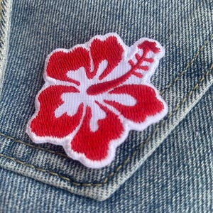 Pode incluir: Patch bordado de flor de hibisco vermelho e branco. O patch está sobre um fundo de jeans azul.
