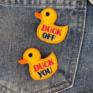 May include: Two yellow duck patches with black eyes and orange beaks are sewn onto a denim pocket. The patches have the phrases "DUCK OFF" and "DUCK YOU" in pink and blue lettering. The denim has a light blue wash.