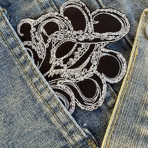 May include: Black octopus patch with white embroidered detailing, set on a denim jacket pocket. The patch shows a detailed design of an octopus with swirling tentacles. The denim jacket is a light blue wash.