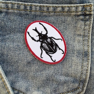 Könnte beinhalten: Ein gestickter Aufnäher mit einem schwarzen Käfer-Design auf einem weißen Oval, umrandet von einem roten Rand. Der Aufnäher ist auf der Tasche eines hellblauen Jeanskleidungsstücks angebracht. Der Käfer hat detaillierte Fühler und Beine.