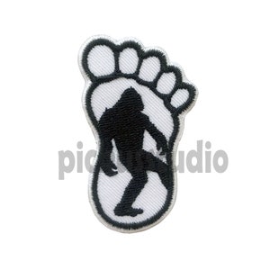 Könnte beinhalten: Weißer gestickter Aufnäher mit einer schwarzen Silhouette eines Bigfoots in einem Fußabdruck.