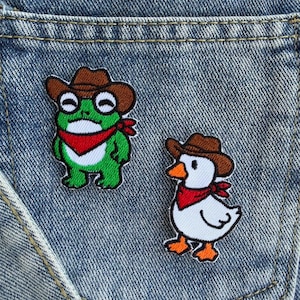 May include: Two embroidered patches featuring a green frog and a white duck, both wearing brown cowboy hats and red bandanas. The frog has a white belly and the duck has orange feet and a beak. These patches are attached to blue denim fabric.