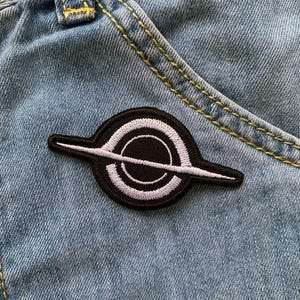 Può includere: Una toppa ricamata in bianco e nero con un cerchio e una linea che lo attraversa, che ricorda un pianeta con un anello. La toppa è cucita su un tessuto di jeans blu.