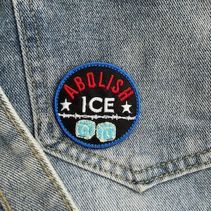 May include: A round patch with the words "ABOLISH ICE" in red and white, with two stars and two blue ice cube graphics. The patch is on a blue denim pocket.