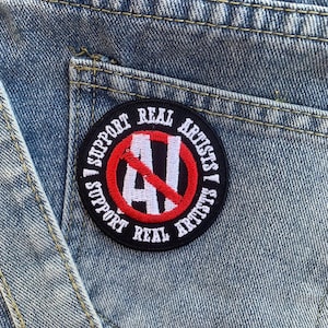 Puede incluir: Un parche bordado en negro y rojo con el texto "Support Real Artists" y un c&iacute;rculo rojo con una l&iacute;nea diagonal que atraviesa la letra "AI" en blanco.