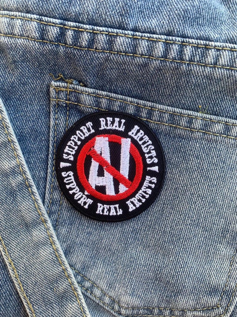 Puede incluir: Un parche bordado en negro y rojo con el texto "Support Real Artists" y un c&iacute;rculo rojo con una l&iacute;nea diagonal que atraviesa la letra "AI" en blanco.