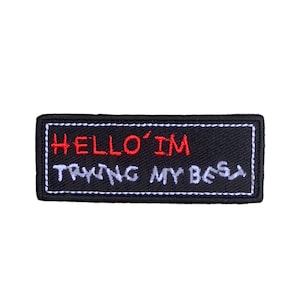 以下が含まれることがあります： 黒地に白のステッチが施された刺繍パッチ。パッチには「HELLO'IM TRYING MY BEST」と赤と白の糸で刺繍されています。