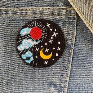 May include: A round, embroidered patch featuring a celestial design. The patch has a black background with a red sun, a yellow crescent moon, white stars, and blue clouds. The patch is sewn onto a denim pocket.