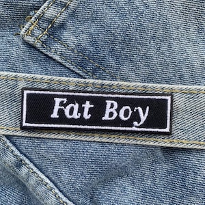 Op de afbeelding: Close-up van blauwe denimstof met een zwart rechthoekig label. Het label heeft witte tekst met de tekst "Fat Boy". De denim heeft zichtbare stiksels en een versleten uiterlijk.