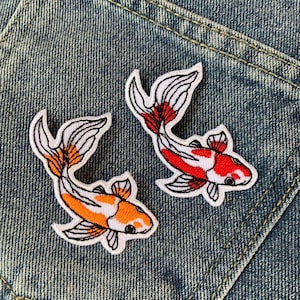 Peut inclure: Deux patchs brodés thermocollants de poissons koï. Les poissons sont blancs avec des accents orange et rouge. Les patchs sont sur un fond en jean bleu.