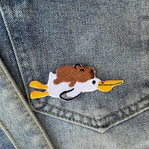 May include: Embroidered patch featuring a white duck with yellow feet and beak, carrying a brown animal on its back. The patch is sewn onto a denim pocket, showcasing a playful design.