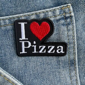 May include: A black embroidered patch with a red heart symbol and the white text I Pizza, featuring a bold design. The patch is attached to a light blue denim fabric surface with visible stitching and a pocket seam detail in the background.