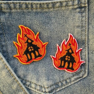 Op de afbeelding: Twee geborduurde patches met een zwart kerk silhouet in oranje en rode vlammen. De patches zijn op een denim zak genaaid en tonen een uniek, gedurfd ontwerp. Perfect om kleding of accessoires een alternatieve stijl te geven.