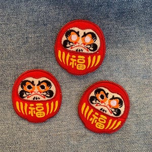 May include: Three red and white Japanese Daruma dolls with black eyes and a yellow "福" character on the front. The dolls are embroidered and are on a blue denim background.
