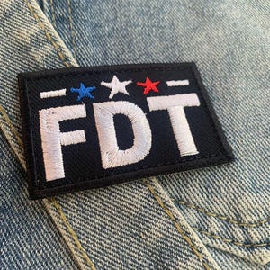 May include: A black patch with white embroidered letters spelling "FDT" and three stars above the letters. The stars are red, white, and blue.