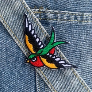 Puede incluir: Un parche bordado colorido de un pájaro golondrina con detalles rojos, amarillos, verdes y negros. El parche está sobre un fondo de mezclilla azul.