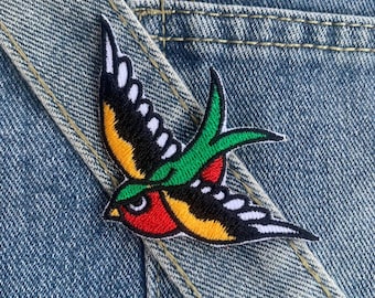 Puede incluir: Un parche bordado colorido de un pájaro golondrina con detalles rojos, amarillos, verdes y negros. El parche está sobre un fondo de mezclilla azul.