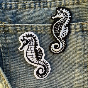 Può includere: Due toppe ricamate a forma di cavalluccio marino, in bianco e nero, su sfondo denim. Le toppe presentano cuciture dettagliate, con un corpo nero e un contorno bianco. Sono probabilmente destinate all'abbigliamento o agli accessori.