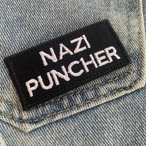 Könnte beinhalten: Ein schwarzer Patch mit weiß gesticktem Text, der "NAZI PUNCHER" auf einem blauen Jeansstoff-Hintergrund lautet.