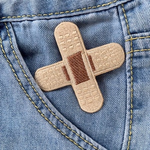 Op de afbeelding: Een beige stoffen verband met een bruin middenstuk zit in de zak van een blauwe spijkerbroek.