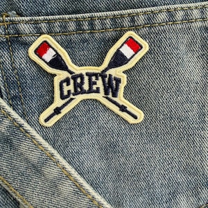 May include: A blue denim fabric background featuring an embroidered patch. The patch displays two crossed oars with navy, white, and red blades, centred with the word CREW in bold navy blue letters on a cream-coloured background with a stitched border.
