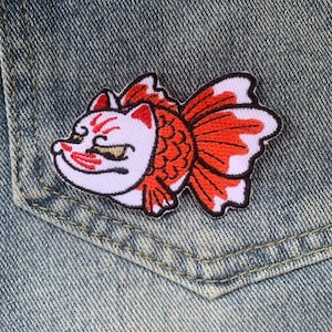 May include: Embroidered patch featuring a stylized goldfish design in red, white, and black. The patch is placed on a denim pocket, showcasing the patch's details and colors. The goldfish has a detailed face and flowing fins.