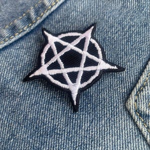 Può includere: Toppa ricamata in bianco e nero con un design a pentagramma. La toppa è cucita su un tessuto denim blu, con cuciture visibili. Il pentagramma bianco è delineato in nero su sfondo nero.