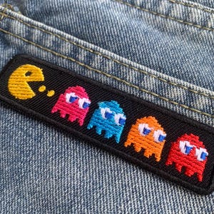 Puede incluir: Parche bordado con el personaje de Pac-Man y cuatro fantasmas en rosa, azul, naranja y rojo. El parche tiene un fondo negro y está cosido sobre tela vaquera azul.