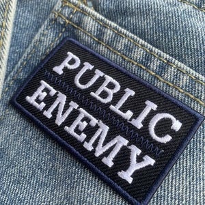 Puede incluir: Un parche rectangular con las palabras "PUBLIC ENEMY" bordadas en blanco sobre un fondo negro. El parche está cosido sobre una tela vaquera azul claro, con costuras visibles y un borde azul oscuro.