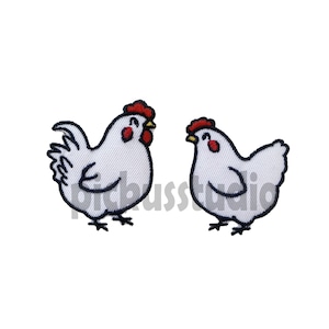 Könnte beinhalten: Zwei weiße gestickte Hühner, die sich gegenüberstehen. Die Hühner haben rote Kämme und Kehllappen und schwarze Augen.