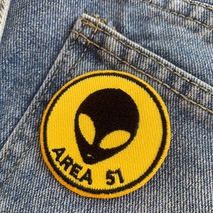 May include: A circular yellow patch featuring a black alien head and the words "AREA 51" in black. The patch is attached to blue denim fabric, possibly a garment.