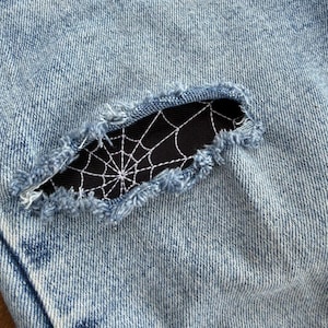 May include: Light blue acid-wash denim fabric featuring a horizontal distressed rip. The opening reveals a black fabric patch underneath, which is embroidered with a white spiderweb design featuring thin, radiating lines and concentric circles.