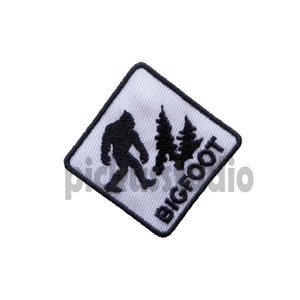 Könnte beinhalten: Weißer quadratischer Patch mit einem schwarzen gestickten Bild von Bigfoot, der mit einem Kiefernbaum hinter sich geht. Der Text "BIGFOOT" ist in Schwarz unter dem Bild gestickt.