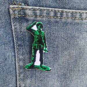 May include: A green plastic toy soldier patch with white details, saluting, on a blue denim background.