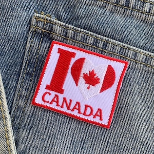 Puede incluir: Un parche rectangular con el texto "I CANADA" en letras rojas y un corazón rojo con una hoja de arce blanca sobre un fondo blanco. El parche está cosido en un bolsillo de mezclilla.