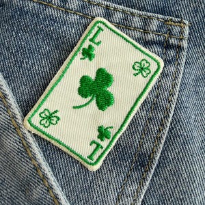 May include: A rectangular embroidered patch shaped like a playing card with a white background and green border. The patch features a large green shamrock in the centre, smaller shamrocks in the corners, and the letter L at the top and bottom.