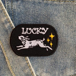 Op de afbeelding: Zwarte geborduurde patch met een wit konijn dat springt en drie gouden sterren. De patch heeft de tekst "LUCKY" in witte letters.