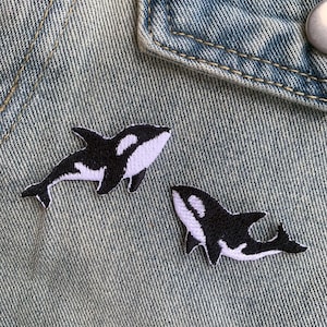 Könnte beinhalten: Zwei gestickte Aufnäher in Form von Orcas, in Schwarz und Weiß, auf einer Jeansjacke. Die Orcas haben weiße Bauch- und Augenpartien und schwarze Körper. Die Jeansjacke ist hellblau.