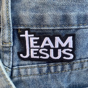 May include: A black and white embroidered patch with the words "Team Jesus" on a pair of blue jeans.