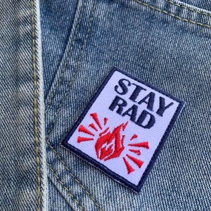 Könnte beinhalten: Nahaufnahme einer Jeansjackentasche mit einem rechteckigen Aufnäher. Der Aufnäher trägt die Worte "STAY RAD" in schwarzer Schrift und ein rotes Flammendesign. Der Aufnäher ist mit einem marineblauen Rand versehen.