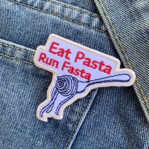 Puede incluir: Parche bordado con el texto "Eat Pasta Run Fasta" en letras rojas sobre fondo blanco. El parche presenta un diseño de espaguetis y tenedor azules. El parche está sobre un fondo de mezclilla.